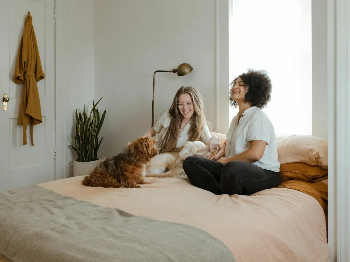 Two women sitting on a bed with a small brown dog in a bright bedroom